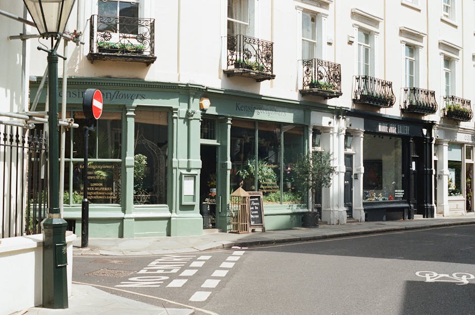 View of a flower shop named 'Kensington Flowers' on a city street corner, featuring large display windows filled with a variety of fresh floral arrangements. The shop's exterior is painted in a muted 