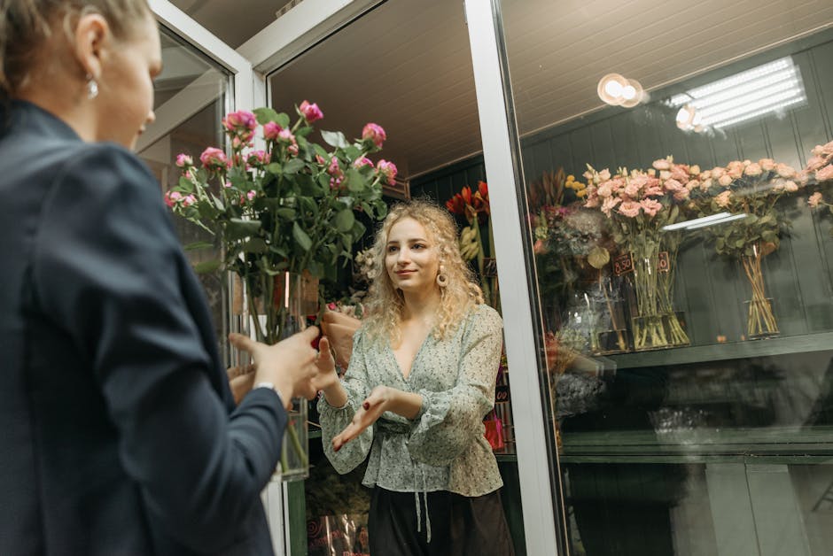 A florist in a dark blazer hands over a large bouquet of pink roses and greenery to a customer inside a floral shop. The bouquet is wrapped in brown paper and held upright, with vibrant pink roses and