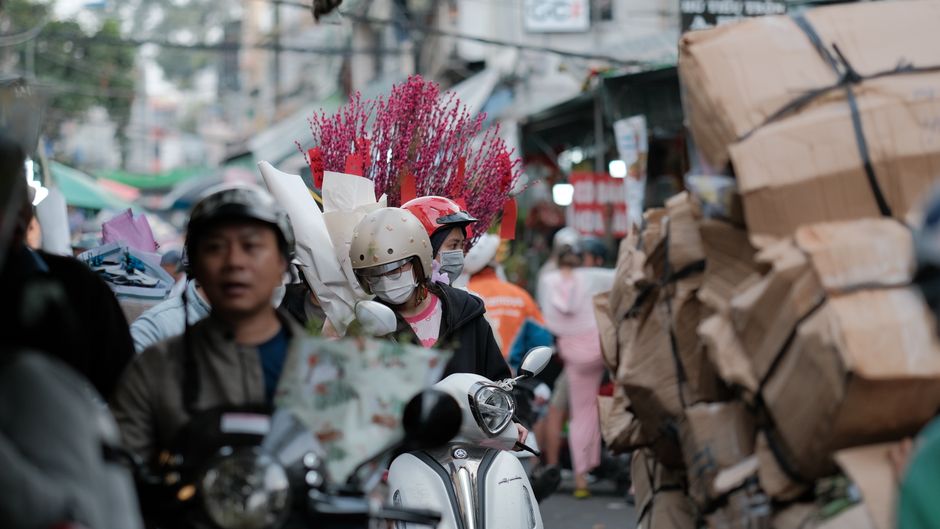 A busy street scene in an Asian market area with numerous motorbikes, some carrying fresh flowers wrapped in white paper, including vibrant pink and red blooms, and others carrying packages or pedestr