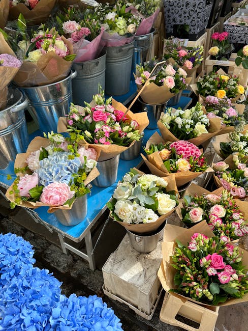 A display of freshly arranged flower bouquets and potted floral arrangements on a table at a flower market. The bouquets feature roses, hydrangeas, and other blooms in soft pink, white, and blue color