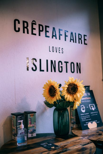 A floral arrangement featuring three large, vibrant yellow sunflowers with dark brown centers, placed in a tall, dark green glass vase on a wooden table. The arrangement is set against a white wall wi