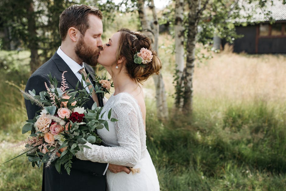 A newlywed couple sharing a kiss outdoors, with the bride holding a large, fresh floral bouquet featuring pink roses, deep red blooms, and greenery, wrapped in light pink paper with a pink ribbon. The