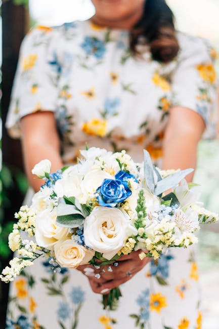 A person in a floral dress holding a bouquet of fresh flowers, featuring white roses, blue roses, and silvery foliage. The bouquet is arranged with a mix of white, cream, and blue blooms, complemented