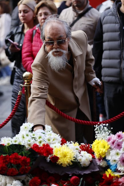 An elderly man with a white beard and glasses is reaching out to place a floral arrangement on a memorial display at an outdoor event. The bouquet contains a vibrant mix of red, white, yellow, and pin